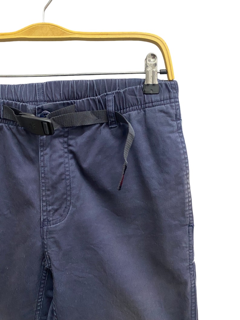 May include: Navy blue trousers featuring an elasticized waistband and an integrated black nylon webbing belt with a plastic side-release buckle. The pants include a front fly detail and are suspended from a light-colored wooden clothes hanger.