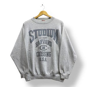 以下が含まれることがあります： 「STUDIUM BIG CHAMP HARVARD CROSSING U.S.A.」の文字がダークブルーでプリントされたヘザーグレーのスウェットシャツ。白い背景に、木製のハンガーに吊るされています。