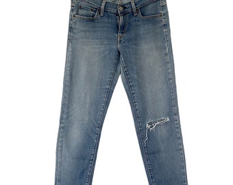 May include: A pair of medium-wash blue denim jeans featuring a classic five-pocket design, a button and zip fly closure, and a distressed frayed hole on the left knee. The jeans are displayed hanging from a gold-toned metal clothes hanger.