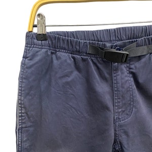 May include: Dark navy blue pants featuring an elasticized waistband with an integrated black nylon webbing belt and a plastic side-release buckle. The pants have a standard fly construction and are displayed hanging from a wooden hanger.