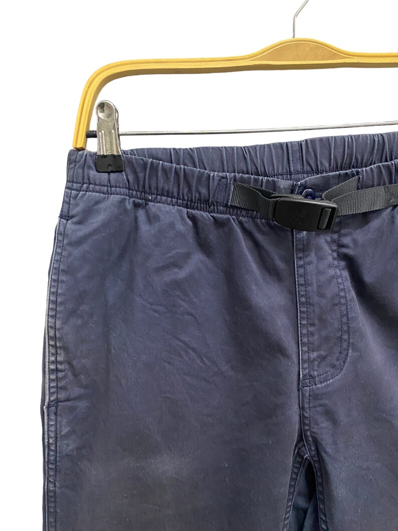 May include: Dark navy blue pants featuring an elasticized waistband with an integrated black nylon webbing belt and a plastic side-release buckle. The pants have a standard fly construction and are displayed hanging from a wooden hanger.
