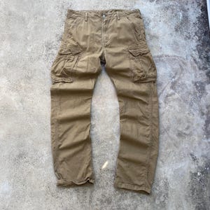 May include: Khaki-colored cargo pants with multiple pockets on the legs and sides. The pants have a button and zipper closure at the waist and are made of a durable fabric. The pants are laid flat on a concrete surface.