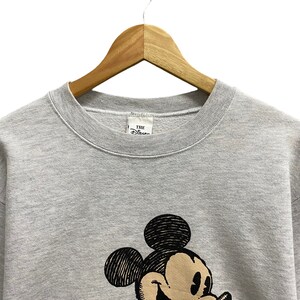May include: A gray sweatshirt featuring a large graphic of Mickey Mouse. The sweatshirt is hanging on a wooden hanger. The Disney label is visible at the neckline. The graphic is in black and tan.
