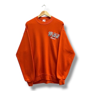 Puede incluir: Sudadera naranja con el texto "WHS Alumni" en escritura blanca y negra. La sudadera tiene cuello redondo y mangas largas. La prenda está colgada de una percha de madera.