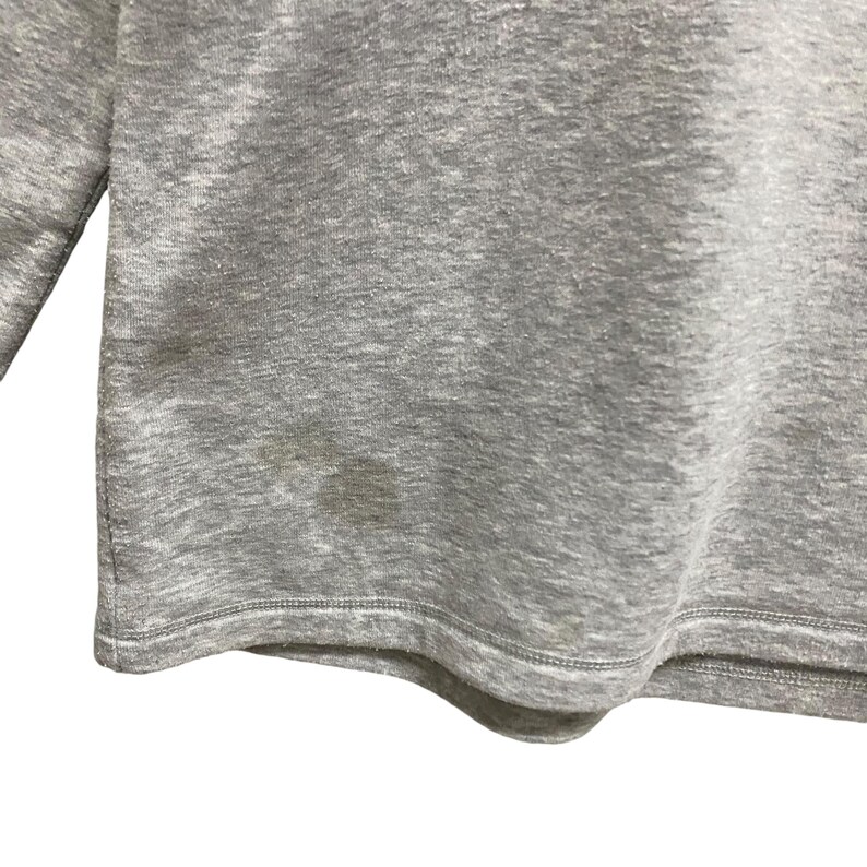 Puede incluir: Una camiseta de manga larga gris jaspeado con cuello redondo. La prenda tiene una mancha en la parte inferior delantera. La tela parece ser un material de punto suave.