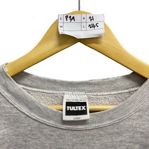 May include: A light gray crewneck sweatshirt featuring a white interior neck label with the black text TULTEX and the size LARGE. A white paper tag is attached to the wooden hanger with the handwritten text C P29, A 21, W, and L 24.5.