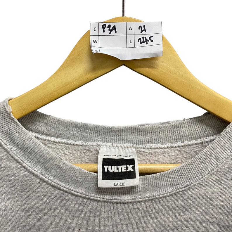 May include: A light gray crewneck sweatshirt featuring a white interior neck label with the black text TULTEX and the size LARGE. A white paper tag is attached to the wooden hanger with the handwritten text C P29, A 21, W, and L 24.5.