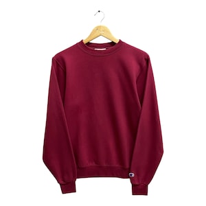 Puede incluir: Una sudadera de cuello redondo de color granate con mangas largas y un pequeño logotipo de Champion en la parte inferior derecha.