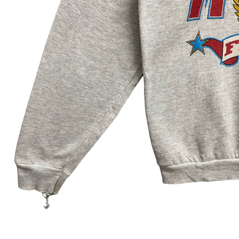 May include: A heather gray long-sleeve sweatshirt featuring a graphic print with red lettering, a yellow laurel wreath, and a blue star. The design includes a red banner with white text. The left sleeve cuff shows visible fraying and loose threads.