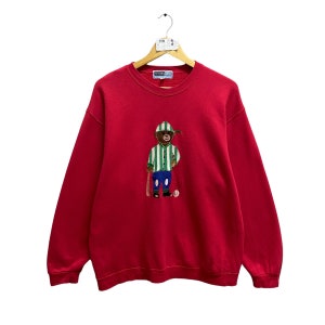 以下が含まれることがあります： 赤いスウェットシャツに、緑と白のストライプの野球帽をかぶってバットを持ったクマが刺繍されています。クマはスウェットシャツの前面に刺繍されています。