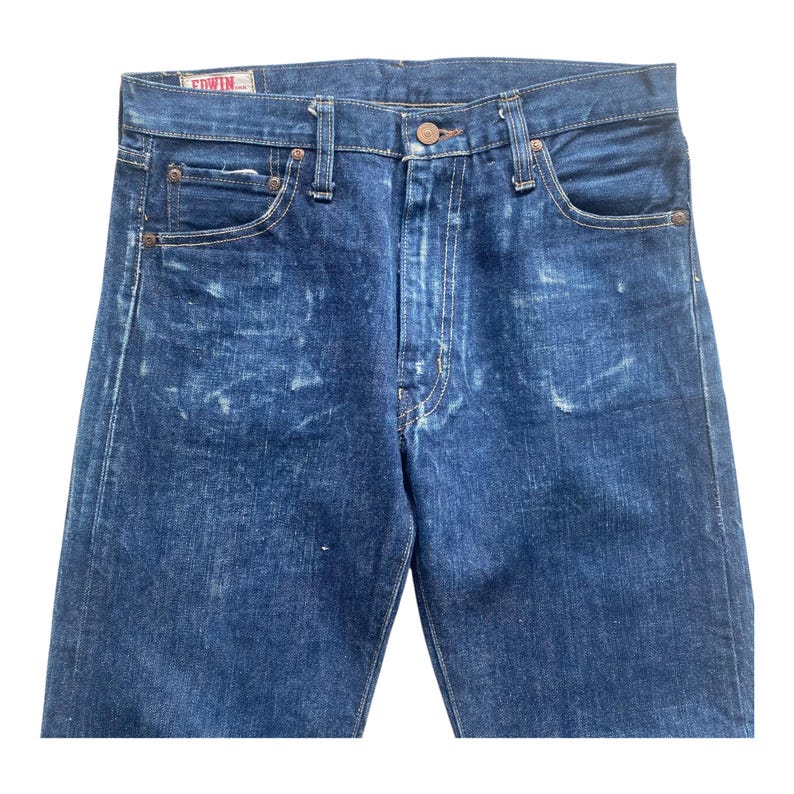 May include: Dark blue denim jeans featuring a faded wash pattern, a button fly closure, and five pockets. The waistband includes belt loops and a small white label with red text reading EDWIN stitched on the interior back of the waistband.