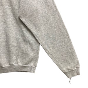 May include: A heather gray long-sleeve sweatshirt featuring a crew neckline and ribbed cuffs. The fabric has a textured, marled appearance. The sleeve cuff shows a small area of fraying with a loose thread hanging from the bottom edge.