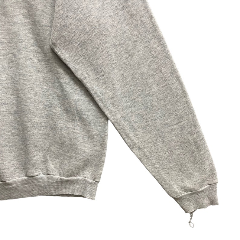 May include: A heather gray long-sleeve sweatshirt featuring a crew neckline and ribbed cuffs. The fabric has a textured, marled appearance. The sleeve cuff shows a small area of fraying with a loose thread hanging from the bottom edge.