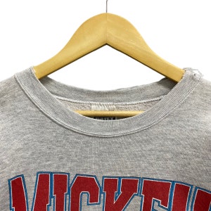 May include: A light gray crewneck sweatshirt featuring a ribbed collar and a partial graphic print in red and blue lettering that reads WICKED. The garment is displayed on a wooden hanger, showing the interior label and fabric texture.