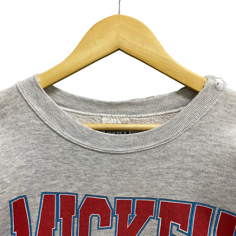 May include: A light gray crewneck sweatshirt featuring a ribbed collar and a partial graphic print in red and blue lettering that reads WICKED. The garment is displayed on a wooden hanger, showing the interior label and fabric texture.