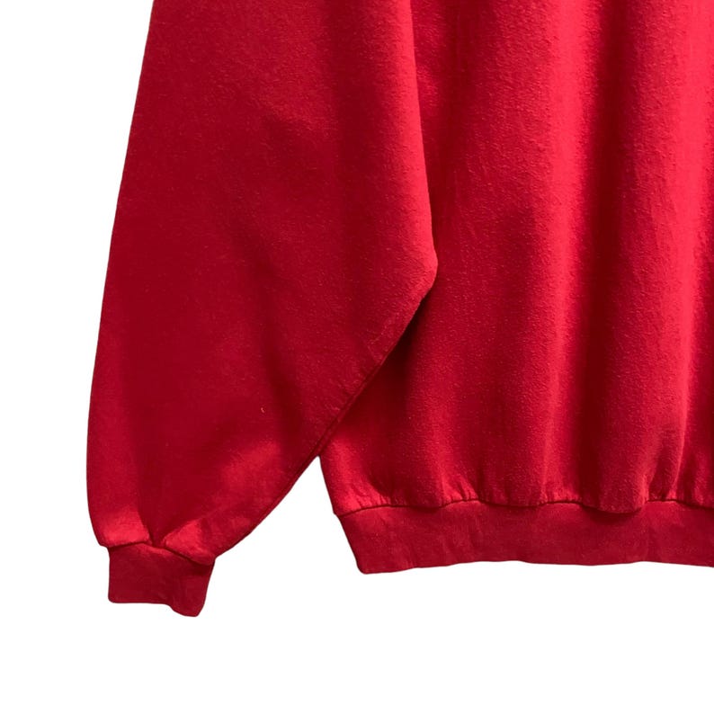 May include: A close-up of a solid red sweatshirt. The image shows the sleeve and part of the body of the garment. The fabric appears to be a soft, knit material. The cuff and hem are ribbed.