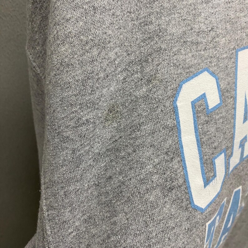 May include: Close-up of a heather gray sweatshirt with the word "CA" in large white letters with a light blue outline. The fabric has a textured appearance, and there is a small stain on the left side of the image.