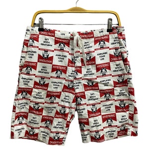 May include: White shorts with a red and black repeating pattern. The pattern includes the word "HYSTERIC" and a graphic of a face. The shorts have a drawstring waist and are hanging on a wooden hanger.