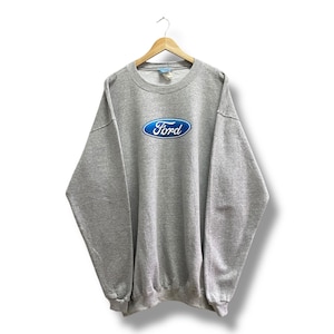 May include: A gray sweatshirt with the Ford logo in blue and white. The crew neck sweatshirt is hanging on a wooden hanger against a white background. The Ford logo is centered on the chest.