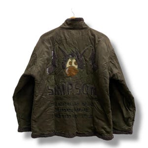 Op de afbeelding: Olijfgroene jas met een cartoonkarakterontwerp op de rug. De jas heeft een bruine fleece voering en het woord "SIMPSON" is geborduurd. De tekst "THE AUSTRALIAN BRAND OR SPORTS APPAREL SINCE 1933 NEW STYLE" is ook zichtbaar.