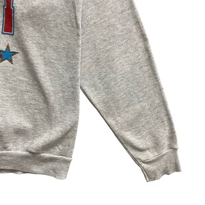 May include: A heather gray long-sleeve sweatshirt featuring a partial graphic print on the left side. The design includes a red vertical element and a blue star with a gold outline. The garment has ribbed cuffs and a ribbed hem at the bottom edge.