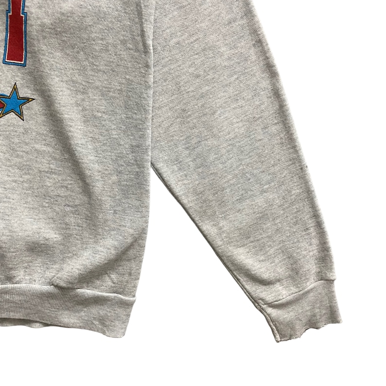 May include: A heather gray long-sleeve sweatshirt featuring a partial graphic print on the left side. The design includes a red vertical element and a blue star with a gold outline. The garment has ribbed cuffs and a ribbed hem at the bottom edge.