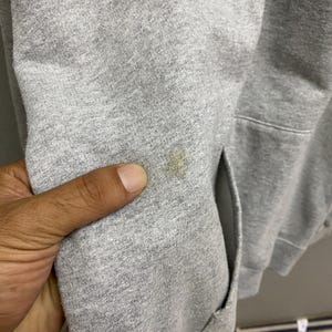 May include: Close-up of a gray hooded sweatshirt with a pocket. The fabric has a stain on the sleeve. The hand of a person is touching the fabric.