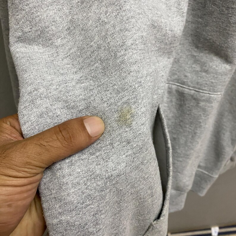 May include: Close-up of a gray hooded sweatshirt with a pocket. The fabric has a stain on the sleeve. The hand of a person is touching the fabric.