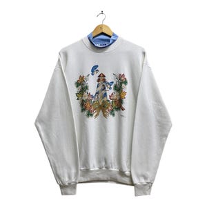 Könnte beinhalten: Weißes Sweatshirt mit blauem Rollkragen. Auf der Vorderseite befindet sich ein Leuchtturm-Design mit blauen Vögeln und einem Blumenkranz. Das Sweatshirt hängt an einem Holzkleiderbügel.