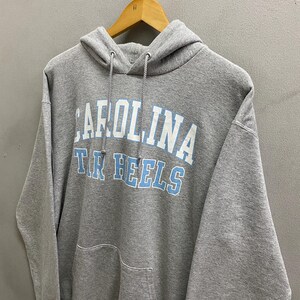 May include: A gray hooded sweatshirt with the text "CAROLINA" above "TAR HEELS" in white and light blue. The sweatshirt has a front pocket and a drawstring hood. The garment is hanging on a wooden hanger.