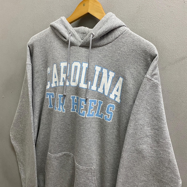 May include: A gray hooded sweatshirt with the text "CAROLINA" above "TAR HEELS" in white and light blue. The sweatshirt has a front pocket and a drawstring hood. The garment is hanging on a wooden hanger.