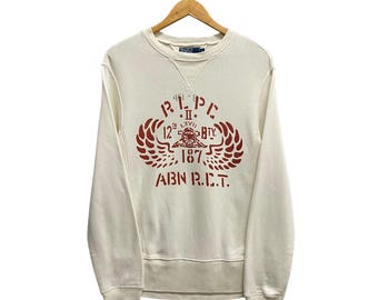 May include: Cream-coloured sweatshirt with a crew neck and long sleeves. The front features a maroon graphic with text and a crest design. The sweatshirt is hanging on a wooden hanger against a white background.
