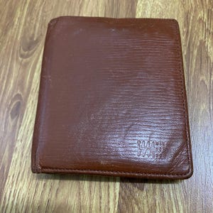 May include: A brown leather wallet with a textured surface. The wallet is rectangular and appears to be closed. The bottom right corner has a logo. The wallet is on a wooden surface.
