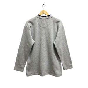 Puede incluir: Una sudadera gris de manga larga con cuello negro y un peque&ntilde;o dise&ntilde;o circular en la espalda. La sudadera est&aacute; colgada de una percha de madera sobre un fondo blanco.