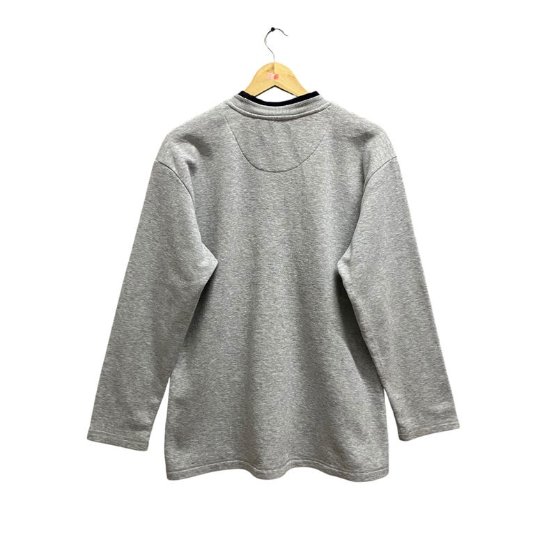 Puede incluir: Una sudadera gris de manga larga con cuello negro y un peque&ntilde;o dise&ntilde;o circular en la espalda. La sudadera est&aacute; colgada de una percha de madera sobre un fondo blanco.