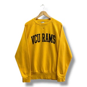 Könnte beinhalten: Senfgelbes Sweatshirt mit dem Text "VCU RAMS" in schwarzen Blockbuchstaben. Das Sweatshirt hat einen Rundhalsausschnitt und lange Ärmel. Das Kleidungsstück hängt an einem Holzbügel.