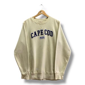 Könnte beinhalten: Cremefarbenes Sweatshirt mit dem Text "CAPE COD MASS" in marineblauen, bogenförmigen Buchstaben. Das Sweatshirt hängt an einem Holzbügel vor weißem Hintergrund. Das Sweatshirt hat einen Rundhalsausschnitt und lange Ärmel.