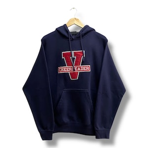 Könnte beinhalten: Marineblauer Kapuzenpullover mit Fronttasche und Kordelzug. Der Pullover hat ein rot-weißes gesticktes Design mit einem großen Buchstaben "V" und dem Wort "CHEERLEADER". Das Kleidungsstück hängt an einem Holzbügel.