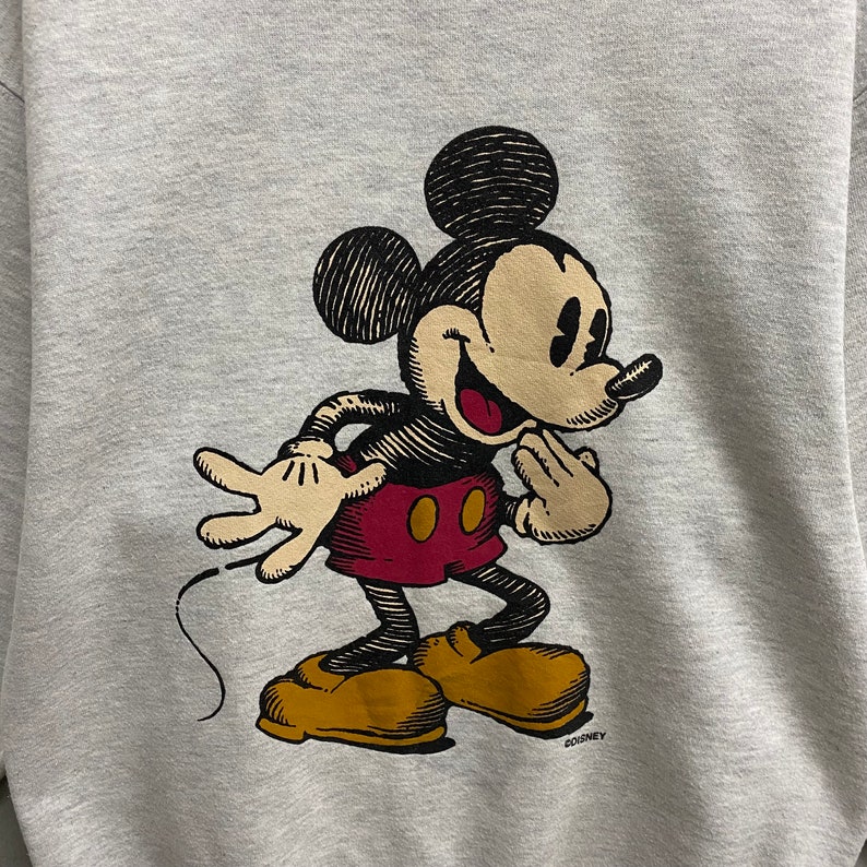 May include: A light gray sweatshirt featuring a vintage illustration of Mickey Mouse. Mickey is depicted in a classic pose, with a smiling expression, red shorts, yellow shoes, and black and white details. The Disney copyright is visible.
