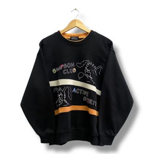 Puede incluir: Sudadera negra con el texto "SIMPSON CLUB ACTIVE SPIRIT" y diseños de personajes de dibujos animados. La sudadera tiene un diseño de rayas blancas y naranjas en el pecho. El cuello es naranja.