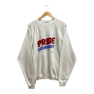 May include: A white sweatshirt with the words "SOFTSEL PRIDE WORLDWIDE" in red and blue lettering. The sweatshirt is hanging on a wooden hanger against a white background. The raglan sleeves are visible.