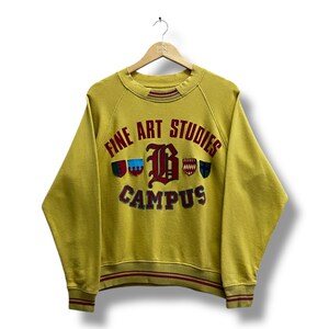 May include: Mustard yellow sweatshirt with the text "FINE ART STUDIES CAMPUS" in red, blue, and green. The sweatshirt has a crew neck and red stripes on the cuffs and hem. The vintage-style sweatshirt is hanging on a wooden hanger.