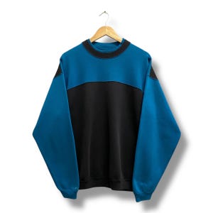 May include: A two-tone sweatshirt in teal and black, hanging on a wooden hanger. The top portion and sleeves are teal, while the lower section is black. The neckline and seams are accented with black stitching.