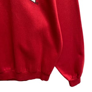 May include: A close-up of a red sweatshirt. The sweatshirt has a crew neck and long sleeves with ribbed cuffs. The fabric appears to be a soft, slightly textured material. The image is well-lit, highlighting the vibrant red color.
