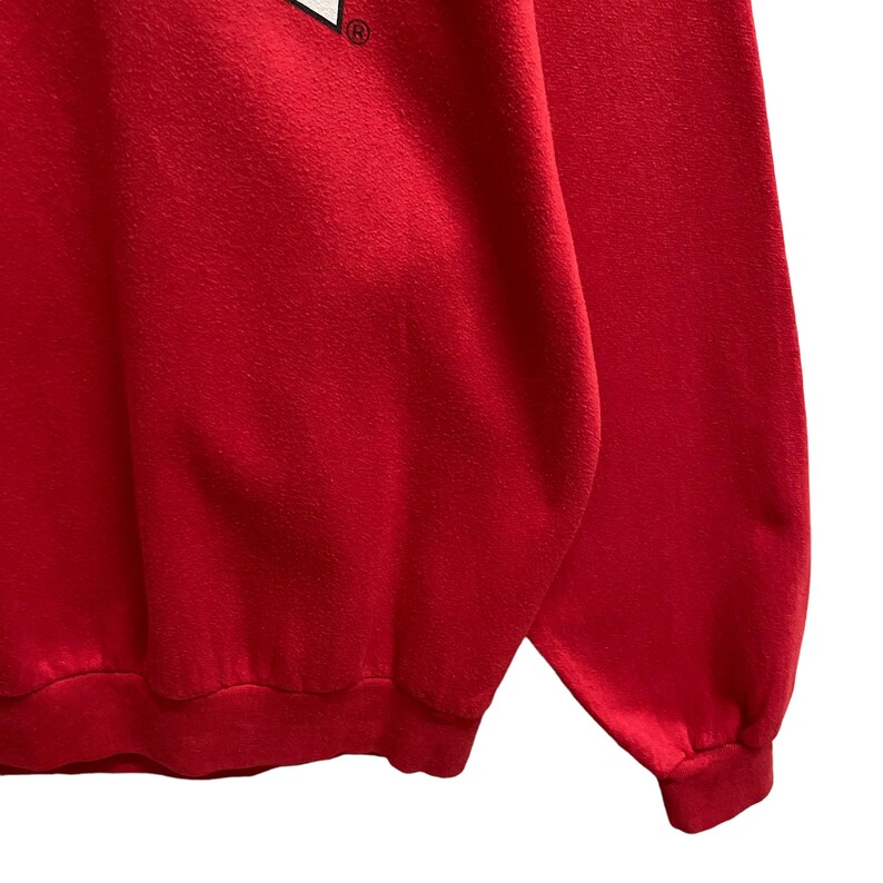 May include: A close-up of a red sweatshirt. The sweatshirt has a crew neck and long sleeves with ribbed cuffs. The fabric appears to be a soft, slightly textured material. The image is well-lit, highlighting the vibrant red color.