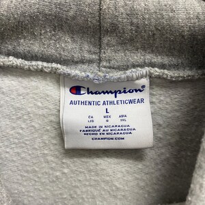 May include: Close-up of a gray Champion hoodie tag. The tag reads "AUTHENTIC ATHLETICWEAR" with the Champion logo. It includes size information and says "MADE IN NICARAGUA" and "CHAMPION.COM".