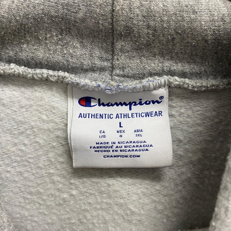 May include: Close-up of a gray Champion hoodie tag. The tag reads "AUTHENTIC ATHLETICWEAR" with the Champion logo. It includes size information and says "MADE IN NICARAGUA" and "CHAMPION.COM".