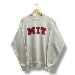 Könnte beinhalten: Ein hellgraues Sweatshirt mit den Buchstaben "MIT" in Burgunderrot mit schwarzer Umrandung auf der Brust. Das Sweatshirt hängt an einem Holzkleiderbügel vor weißem Hintergrund. Es hat einen Rundhalsausschnitt und lange Ärmel.