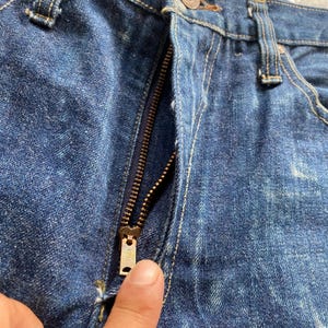 May include: Dark blue denim jeans featuring a brass-toned metal zipper fly closure. The fabric displays a textured weave with visible stitching along the seams and a metallic zipper pull tab centered at the front of the garment.