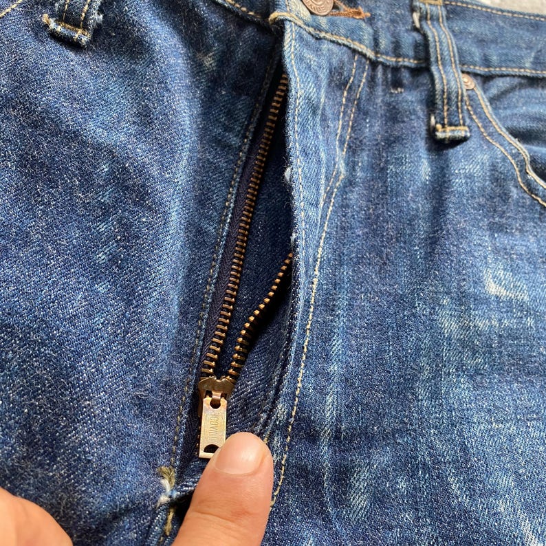 May include: Dark blue denim jeans featuring a brass-toned metal zipper fly closure. The fabric displays a textured weave with visible stitching along the seams and a metallic zipper pull tab centered at the front of the garment.